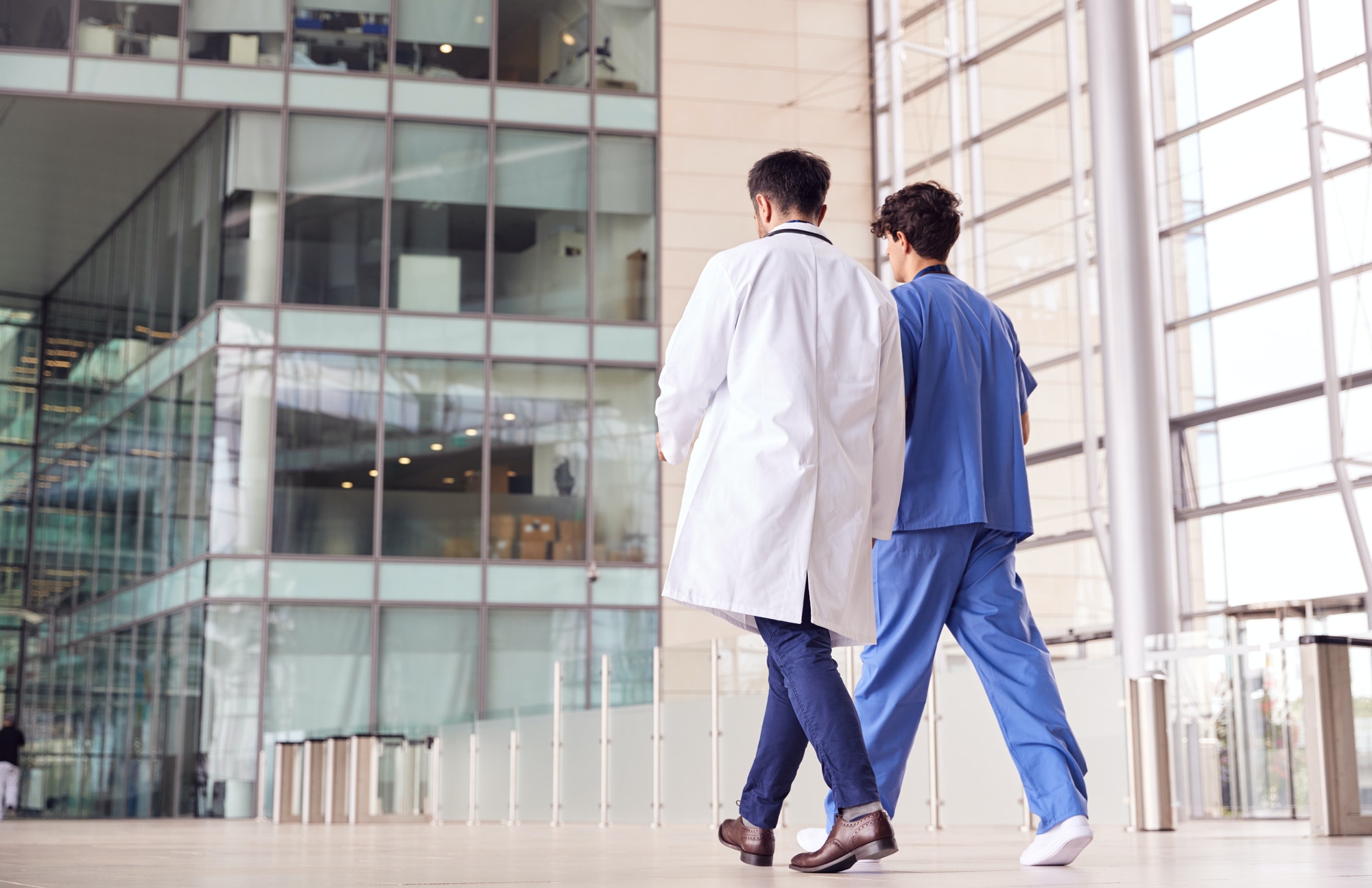 Rear View Of Male Medical Staff Talking As They Walk Through Lobby Of Modern Hospital Building